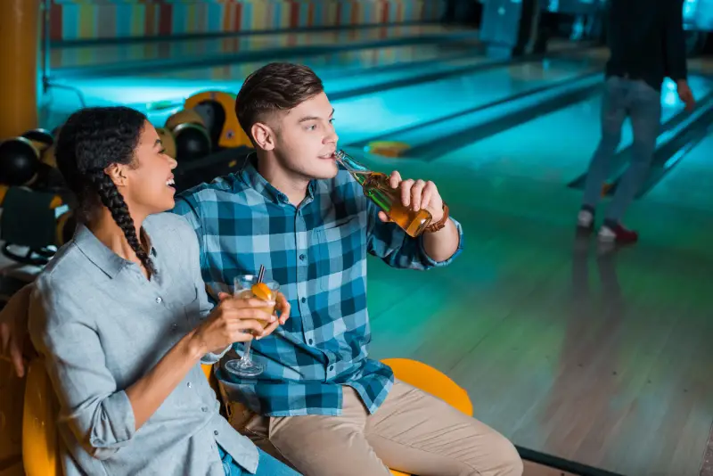 cheerful african american girl holding cocktail glass and talking to boyfriend drinking beer in bowling club